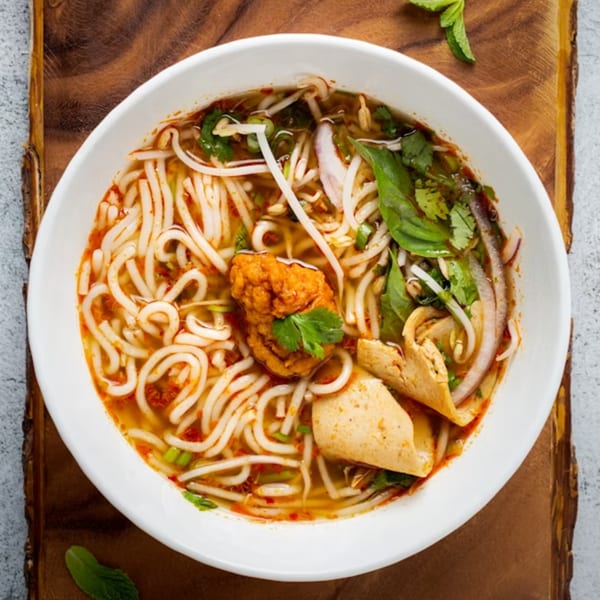 A white bowl of Chinese noodle soup with fried tofu, bean sprouts, and herbs on a wooden board.