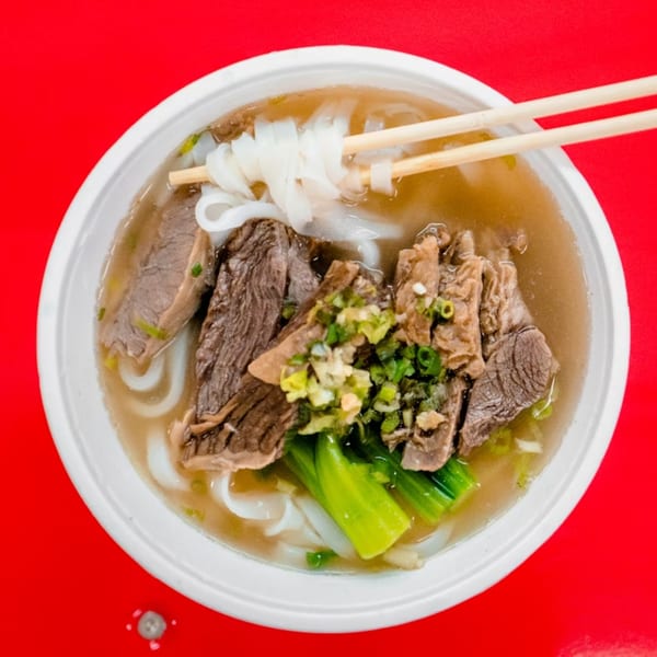 A bowl of Ying Li Restaurant's beef noodle soup with flat noodles, green vegetables, and chopsticks.