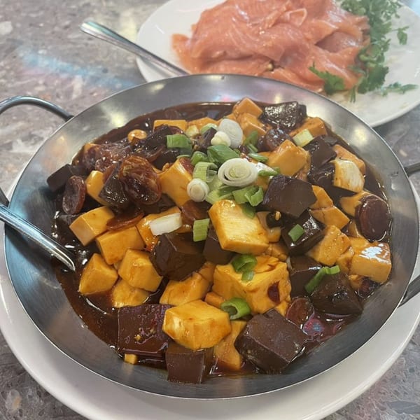 A metal pan of dark braised tofu and mushrooms, garnished with green onions, with salmon in the background.